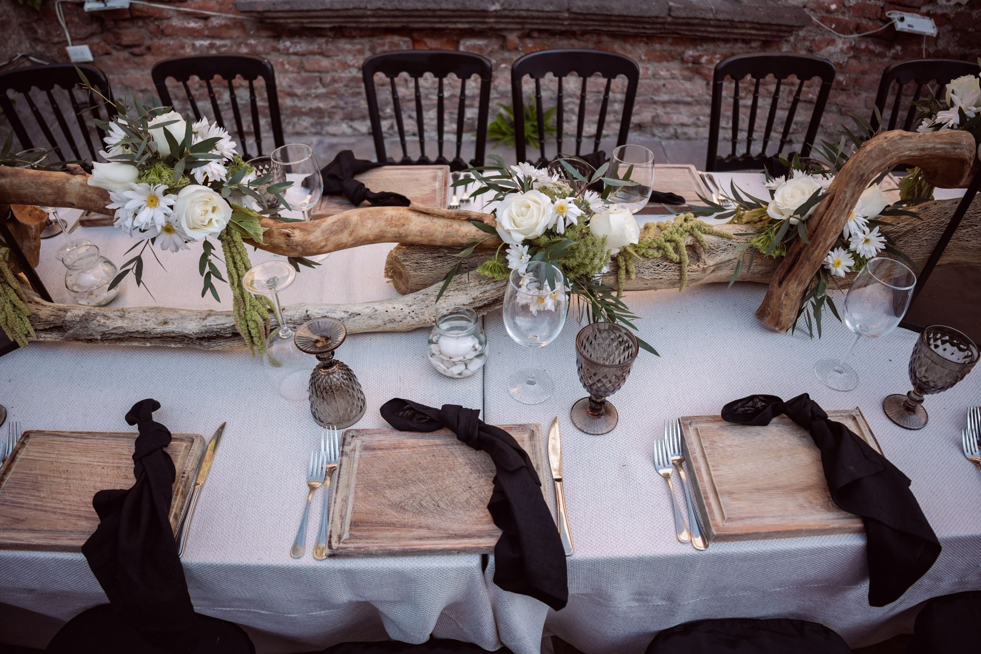 Long courtyard table against an aged brick wall, dressed with black chairs, driftwood and white roses