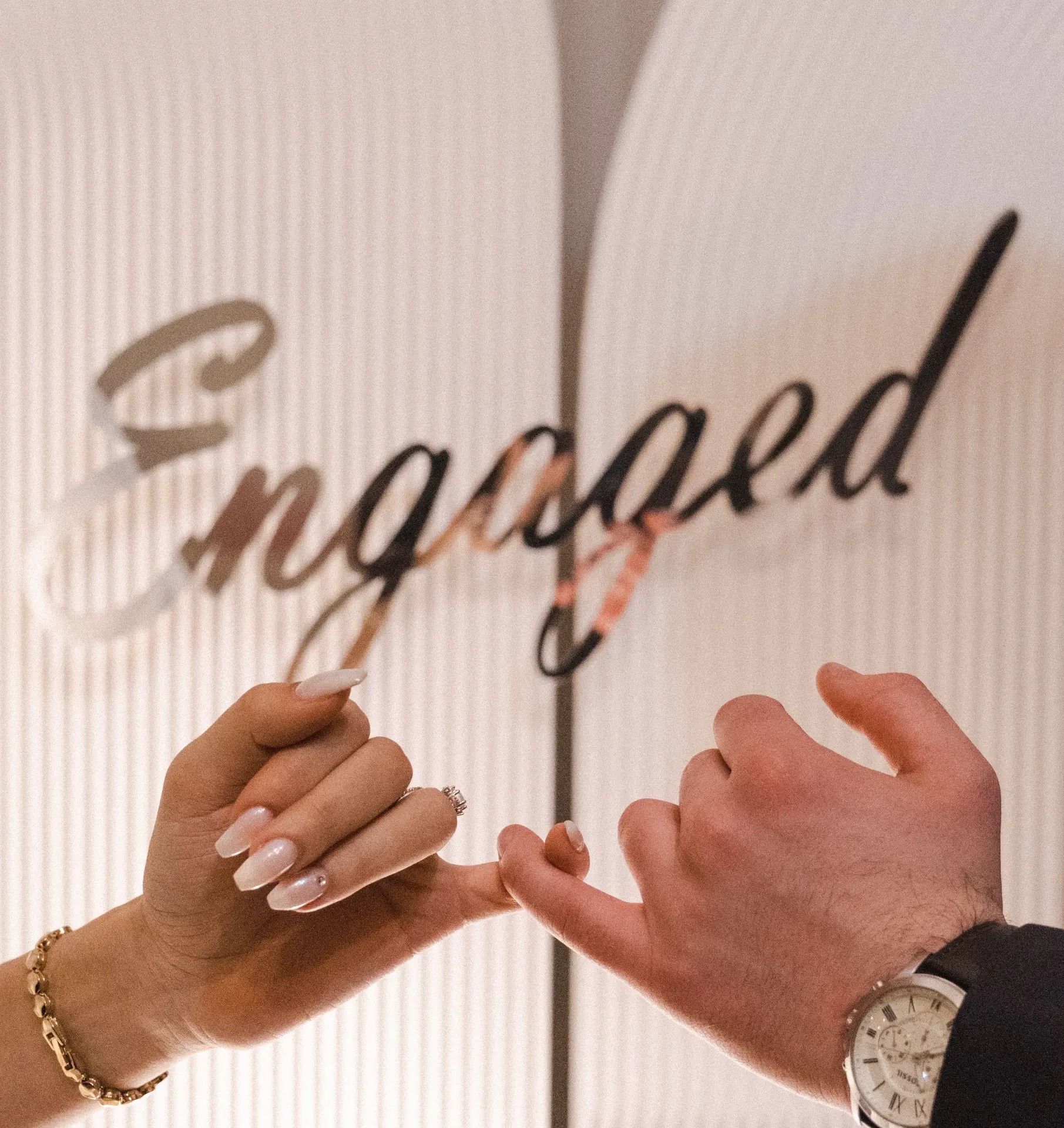 Couple's hands linked in a pinky promise in front of an Engaged sign